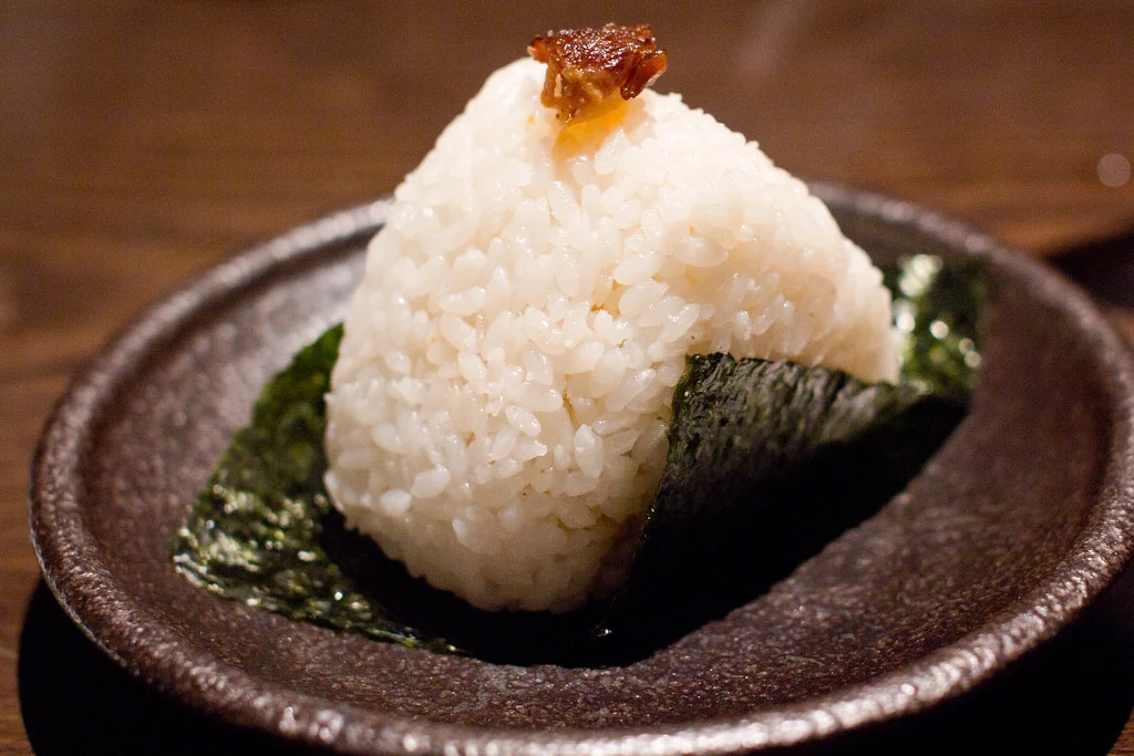 an onigiri sitting on top of a piece of nori with a brown topping on its tip.