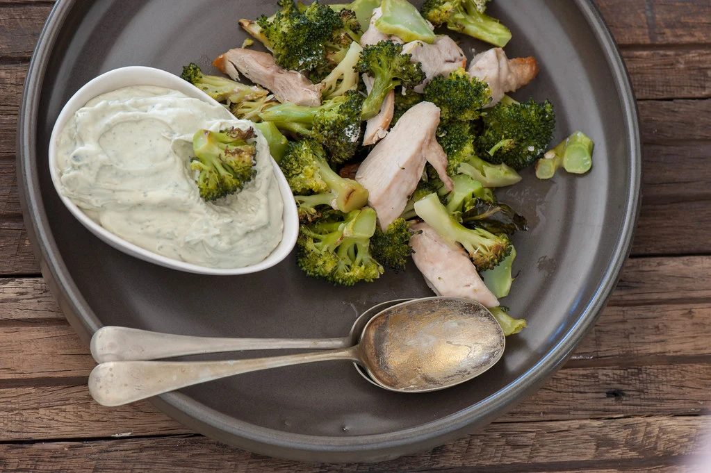 broccoli with some chicken placed next to a bowl of sauce and two spoons in a plate.