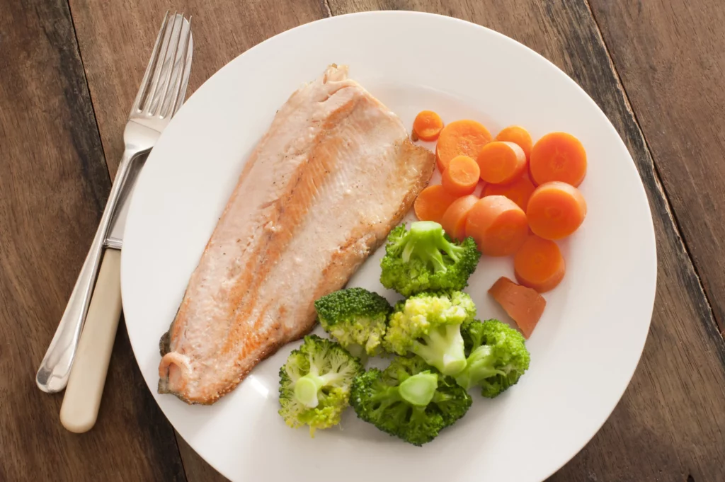 a few pieces of broccoli places next to some slices of carrot and a fish fillet, all in one plate